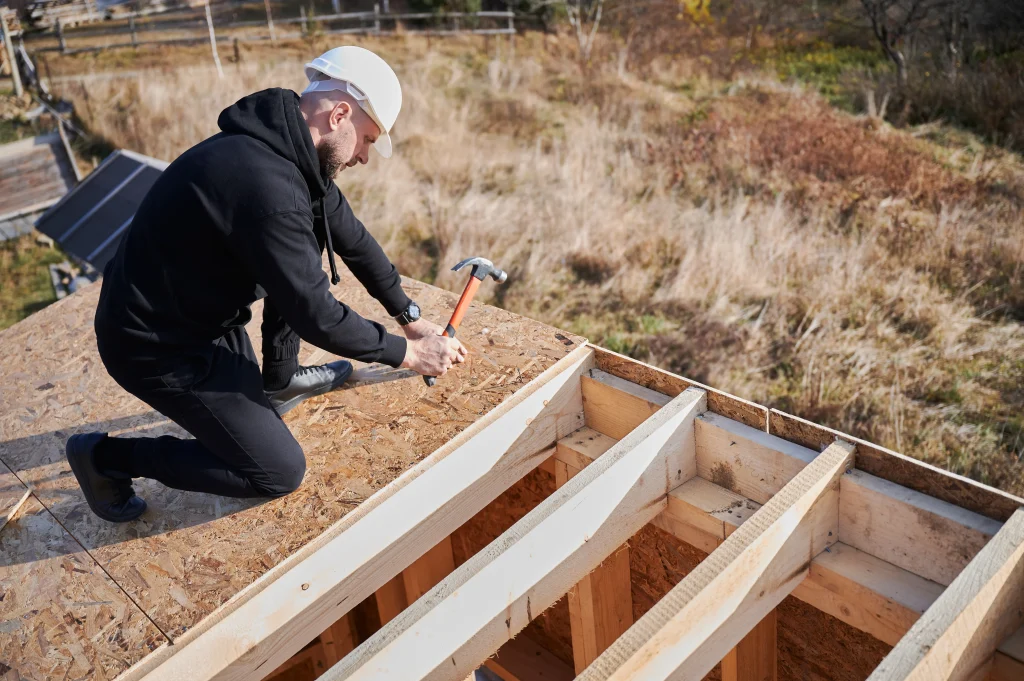 carpenter-hammering-nail-into-osb-panel-while-buil-2025-01-16-22-52-21-utc (1) alt="man in black on knees hand nailing roof decking in spring on half-finished roof"