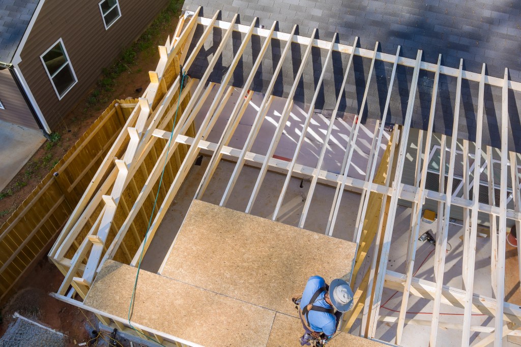 in-course-of-construction-work-carpenter-hammers-2025-03-27-03-43-57-utc (1) (1) alt="man in blue on roof with beams installing roof decking"